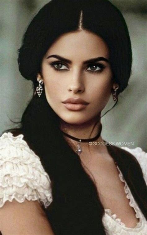 A Woman With Long Black Hair Wearing A White Dress And Earrings On Her Head Is Looking At The Camera