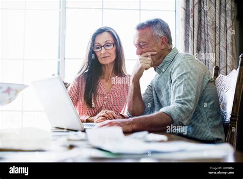 Senior Couple Sitting At A Dining Table Using A Laptop Computer Paperwork And Bills On The