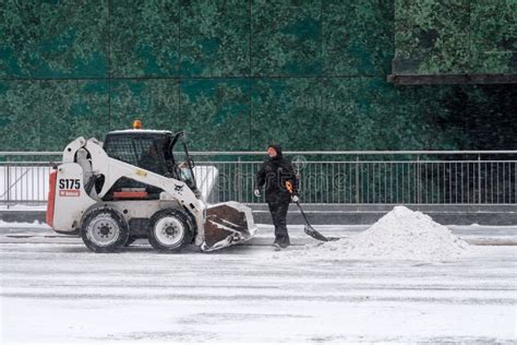 A Utility Worker And A Small Loader Excavator Remove Snow From The Road