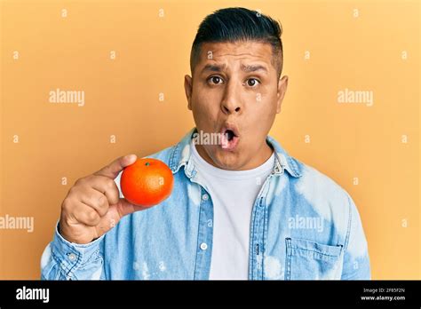 Young Latin Man Holding Tangerine Scared And Amazed With Open Mouth For