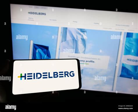 Person Holding Smartphone With Logo Of German Company Heidelberger