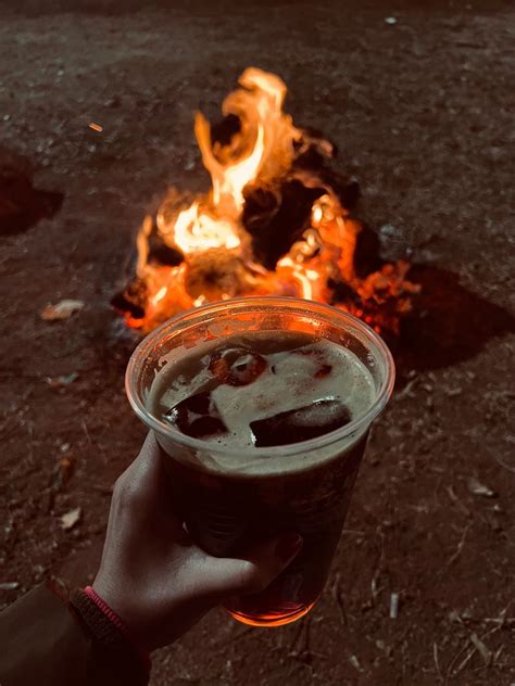 A Person Holding Up A Beer In Front Of A Fire