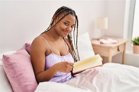 African American Woman Reading Book Sitting On Bed At Bedroom Stock