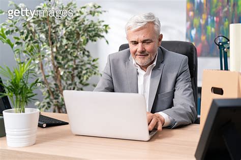 A Mature Man With Gray Hair And Stubble In A Navy Shirt At Desk In The