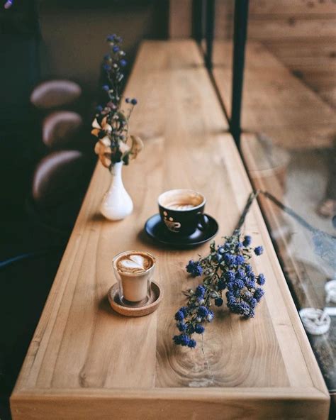 Two Cups Of Coffee Sit On A Wooden Table Next To A Vase With Purple Flowers