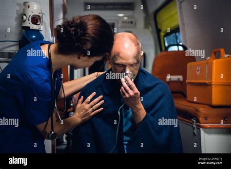 Woman In Blue Medical Uniform Stands With Man Who Sits In Oxygen Mask