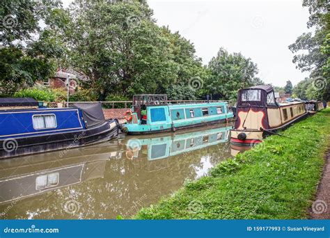 Narrow Boats Moored To Bank Of Canal On Sunny Day With Reflections In