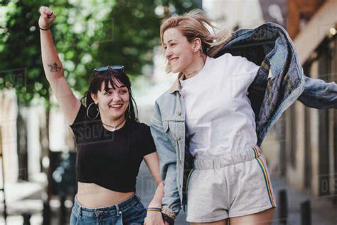 Cheerful Lesbians Holding Hands And Waving Lgbt Flag While Walking On Blurred Background Of City