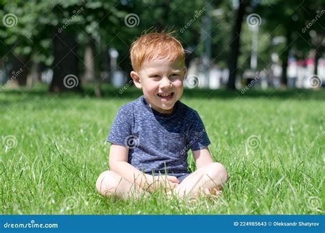 The Red Haired Boy Sits In The Park On The Lawn Under The Bright Sun