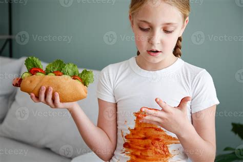The child stained his clothes with ketchup. A girl eats a hot dog