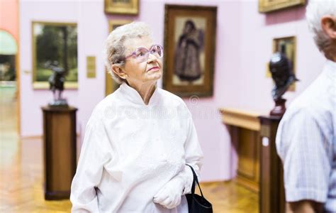 Mature Woman Examines Paintings In An Exhibition In Hall Of An Art