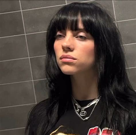 A Woman With Long Black Hair Standing In Front Of A Tile Wall And Wearing A Necklace