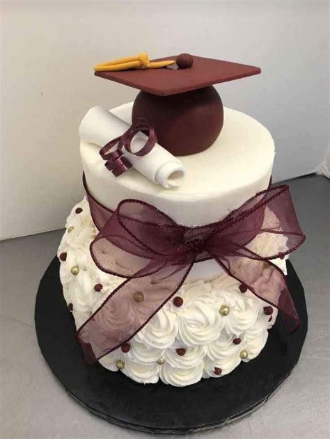 A Graduation Cake Decorated With White Frosting And Brown Ribbon Topped With A Mortar And Diploma