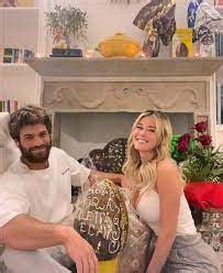 A Man And Woman Sitting On A Couch In Front Of A Fire Place With Flowers