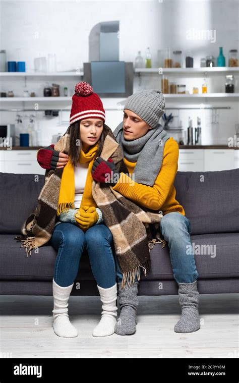 Young Man In Warm Hat And Gloves Covering Freezing Girlfriend With Plaid Blanket In Cold Kitchen