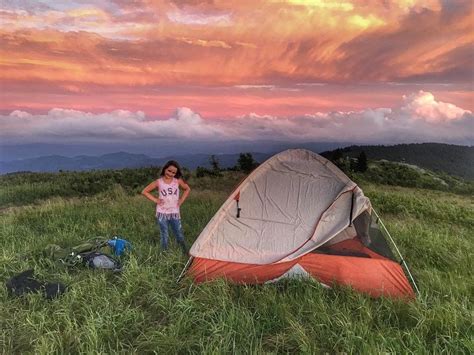 a woman standing next to a tent on top of a lush green hillside under a cloudy sky