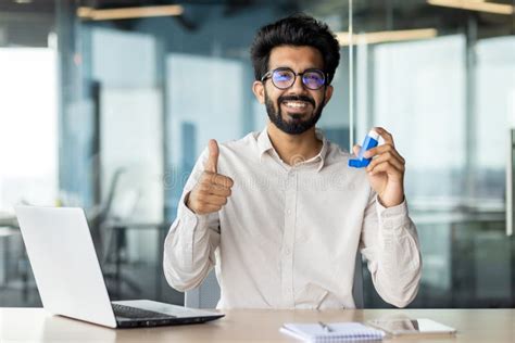 A Man With Glasses And A Beard Smiling While Using A Laptop At A Desk