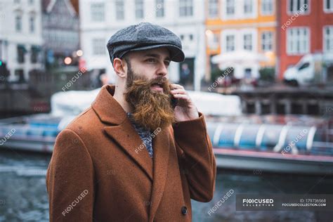 Portrait of bearded man in vintage clothing talking onsmartphone at river in city — young ...