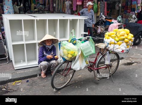 Vietnamese Woman Street Vendor Sitting On The Curb By Her Bicycle Loaded With Mangoes In Ho Chi