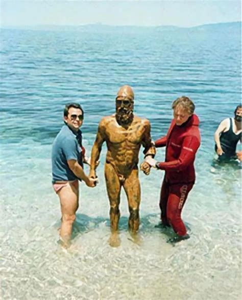 Three People In Wetsuits Standing In The Water With A Statue Of A Man