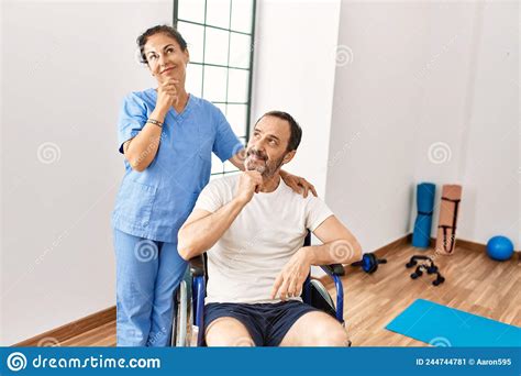 Hispanic Middle Age Man Sitting On Wheelchair And Nurse At Rehabilitation Clinic Serious Face