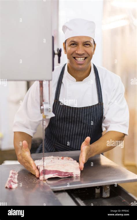 Portrait Of Cheerful Mature Male Butcher Cutting Meat On Bandsaw Stock