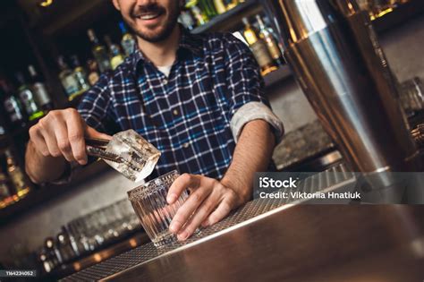 Young Bartender Standing At Bar Counter Putting Ice Cube Into Glass