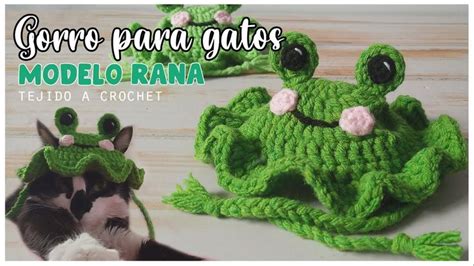 A Green Crocheted Frog Hat With Pink Flowers On It And A Cat Laying