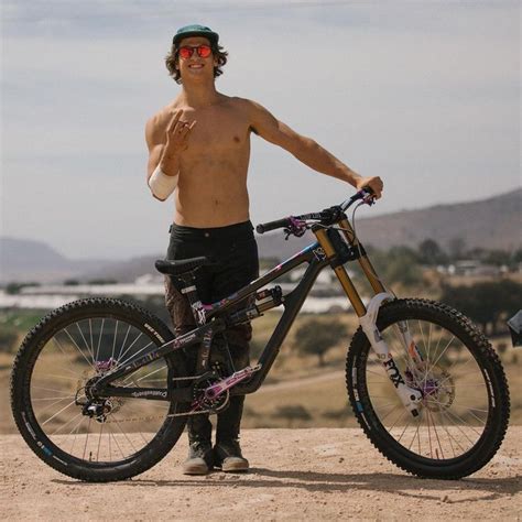 A Shirtless Man Standing Next To His Bike On Top Of A Dirt Field With