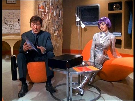 The Man And Woman Are Sitting On Orange Chairs In Front Of A Television