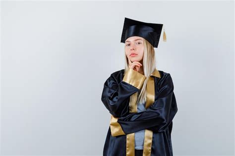 생각 포즈에 서 졸업 가운과 모자에 턱 밑에 손을 넣고 잠겨있는 찾고 금발 소녀 무료 사진