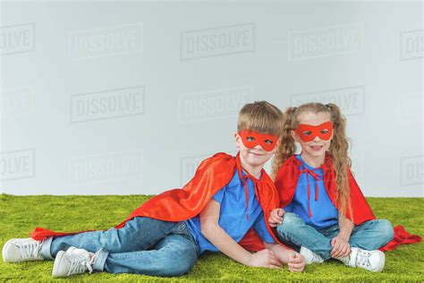 Siblings in superhero costumes sitting on green grass and smiling at camera on grey - Stock ...