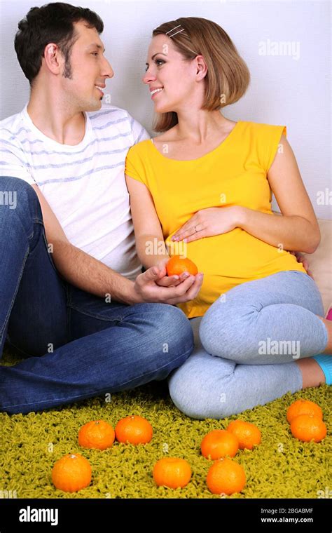 Young Pregnant Woman With Her Husband Sitting On Floor On Gray