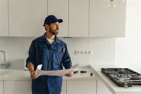 Professional Handyman In Uniform Standing On Kitchen With Home Plan And