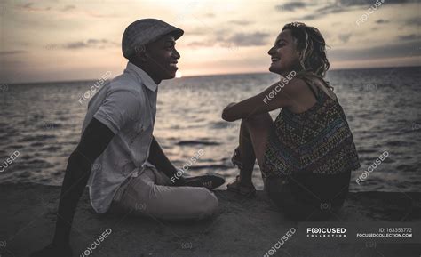 Two People Sitting On Seaside In Front Of Ocean Looking At Each