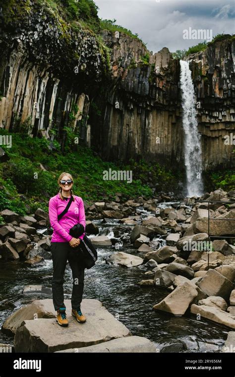 Woman Smiles In Front Of Waterfall Over Basalt Cliff With Greenery