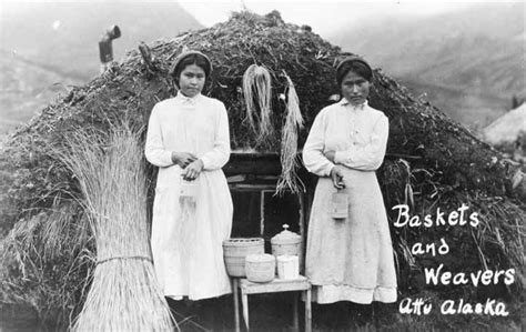 Two Women Standing Next To Each Other In Front Of A Straw Hut With Brooms