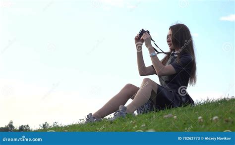 Beautiful Brunette Girl In Black Dress Sitting On The Lawn And Making Photos With Her Camera
