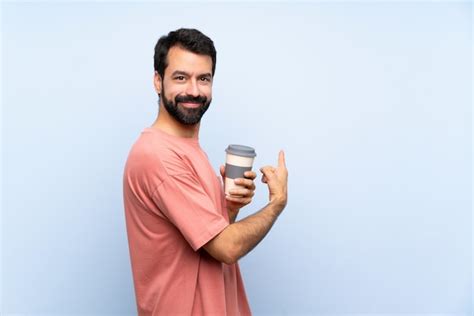 Hombre joven con barba sosteniendo un café para llevar sobre pared azul