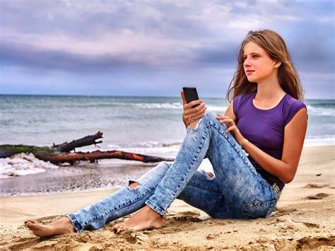 Menina O Telefone Celular Que Senta Se Na Areia Perto Do Mar Foto