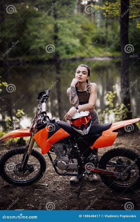 Charming Tattooed Racer Girl Wearing Motocross Outfit With Semi Naked Torso Leaning On Her Bike