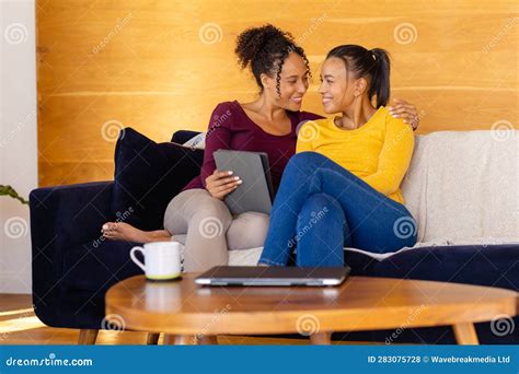 Happy Biracial Lesbian Couple Embracing And Using Tablet On Sofa In