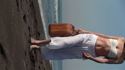 Mature Woman Walking With Brown Suitcase On Black Sandy Beach On