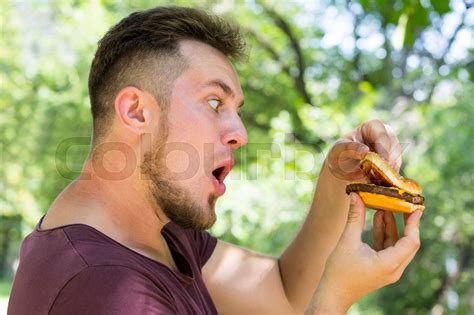 Emotional Young Guy Eating A Cheeseburger On The Nature Stock Image