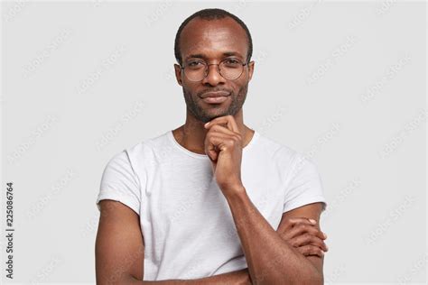 Studio Shot Of Serious Black Man Keeps Hand Under Chin Wears