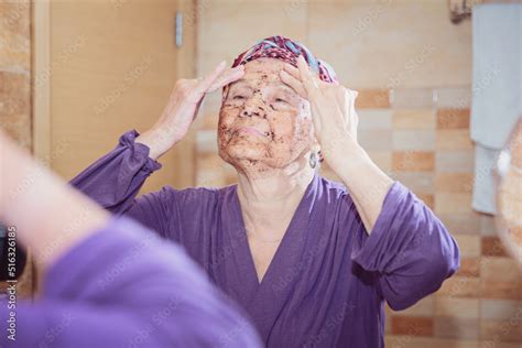 Foto De Mature Woman Standing At The Mirror In The Bathroom And Putting A Scrub On Her Face