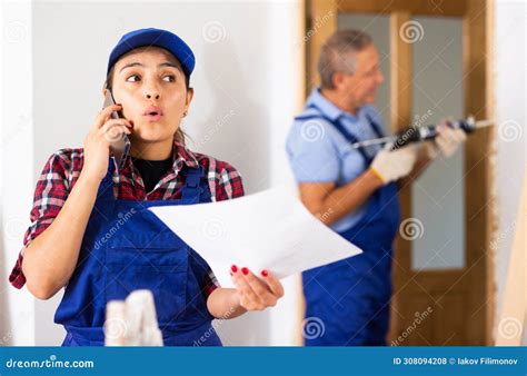 Worried Latin Female Repairer In Uniform Talking On A Mobile Phone While Holding A Work Plan