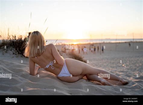 A Blonde Woman Wearing A White Bikini Posing Facing Backwards On The Beach During The Sunset