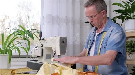 Senior Man Seamstress Sitting And Sews On Sewing Machine In Studio