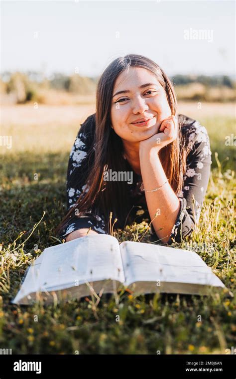 Smiling Christian Girl Lying On The Grass Reading Her Bible In The Field At Sunset Looking At